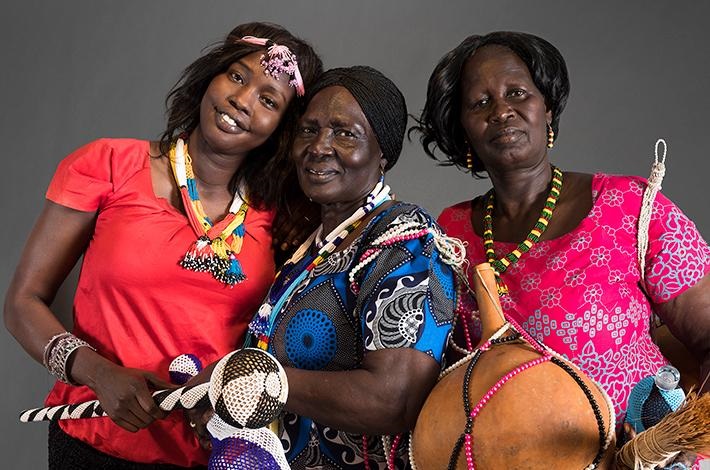 Group of three women wearing traditional Sudanese clothing, smiling and standing closely to one another