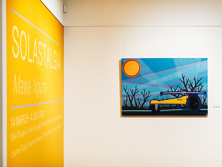 'Solastalgia Mawa Ngurra' written on a bright yellow wall next to an abstract painting of a machine amongst trees under the sun