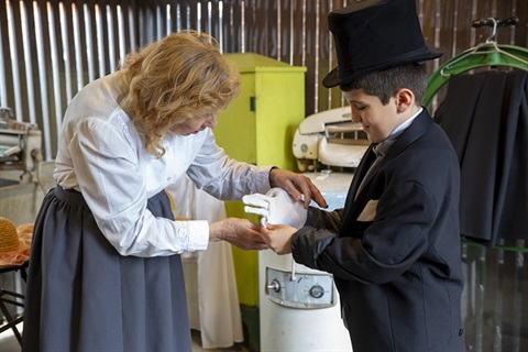 A woman in vintage clothing, helps a child wearing a top hat put on a white glove. 