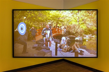 Video still showing people dressed up in a garden projected on a large screen