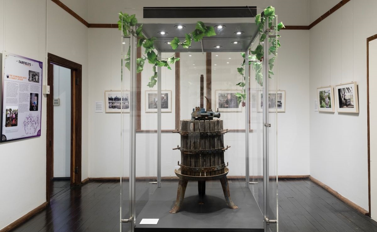 Historical wooden wine press in glass display case in the centre of a room with a gallery of framed photographs