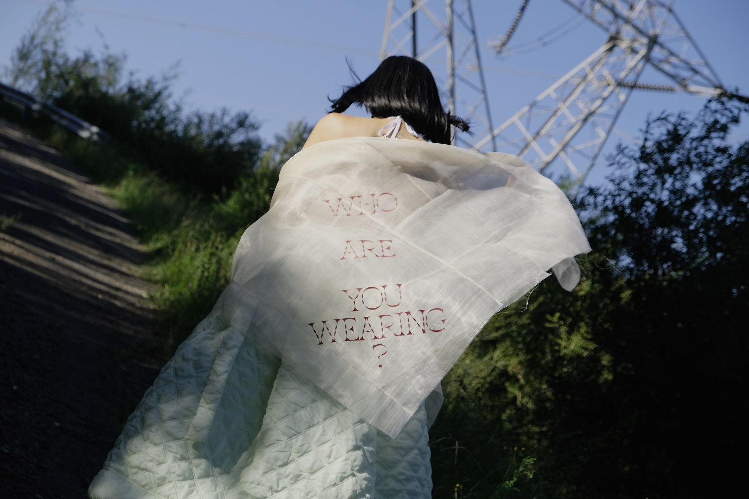 woman wearing a white cape walking up a hill in a park setting. The cape reads 'Who Are You Wearing?'