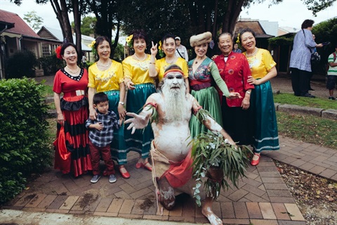 Group of women wearing varying traditional cultural costumes, smiling and posing, with Aboriginal man painted with white ochre and holding a bundle of eucalyptus branches
